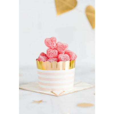 A pink treat cup with white stripes and a gold foil rim, filled with pink heart shaped candies, displayed on a pink and gold napkin on a white surface.