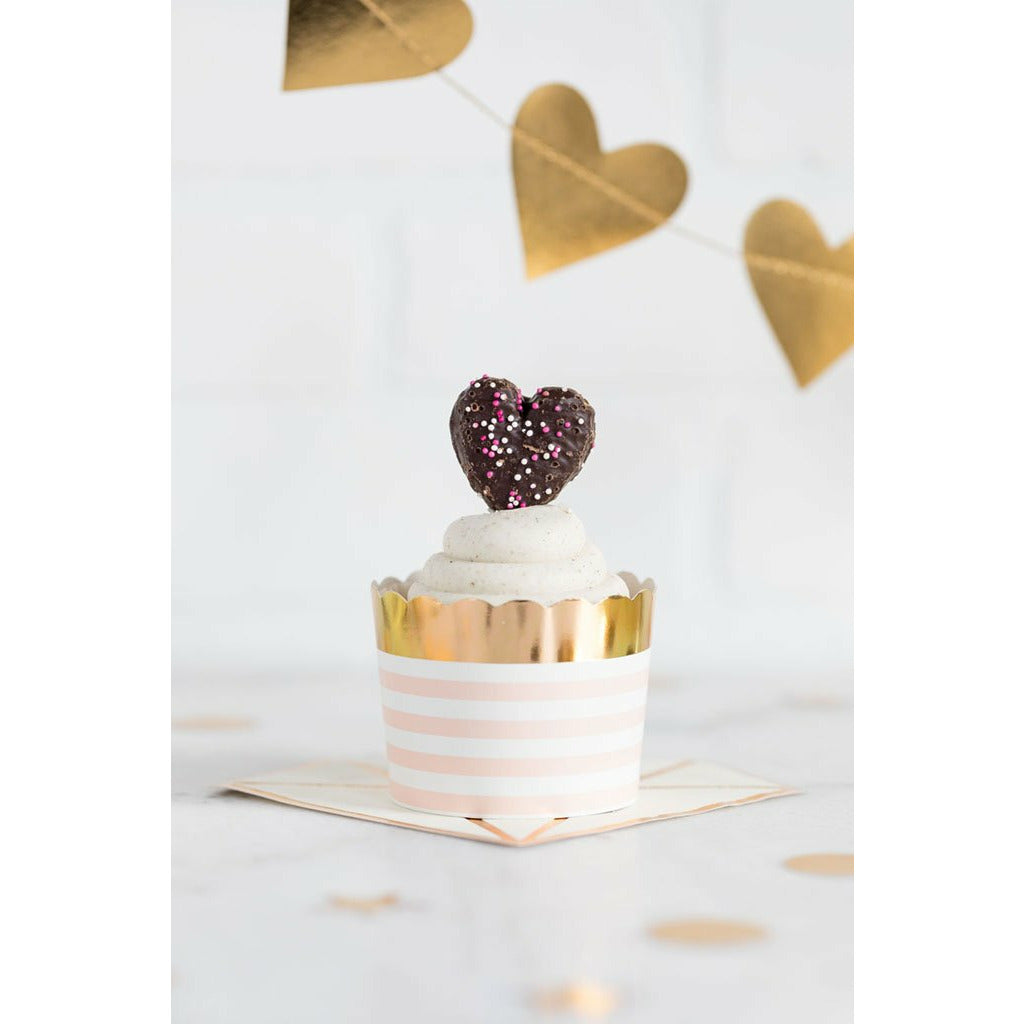A pink treat cup with white stripes and a gold foil rim, filled with white frosted cupcake which is topped with a chocolate covered heart-shaped cookie, displayed on a pink and gold napkin on a white surface with a gold foil heart shaped banner in the background.