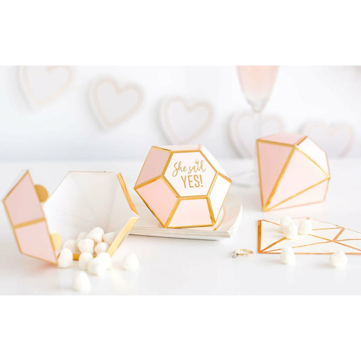 3 diamond-shaped die-cut pink favor boxes with gold foil trim detail and gold foil print that reads "she said yes" in the center. 1 favor box is open with white mint candy pouring out.