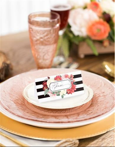 black and white striped place cards with a multi-color floral print surrounding space for names on top of a dinner plate with wine glasses and a floral arrangement in the background.