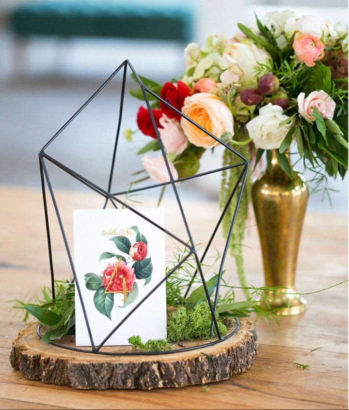 A folded tent style table number card with gold foil accents and a floral print, displayed in a black triangular frame, placed on a wooden platter. A gold vase floral arrangement in background