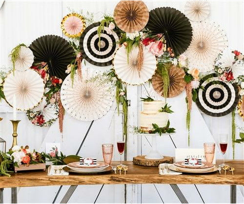 Rustic chic table setting and decorations including a collection of decorative paper fans with various patterns, including floral, polka dots, and stripes, in shades of white, gold, black, red, pink, yellow, and green on a white wall with fern stems placed throughout. Wooden table with wine glasses, candle sticks, a cake on cake stand, and placecards on top of dinner plates