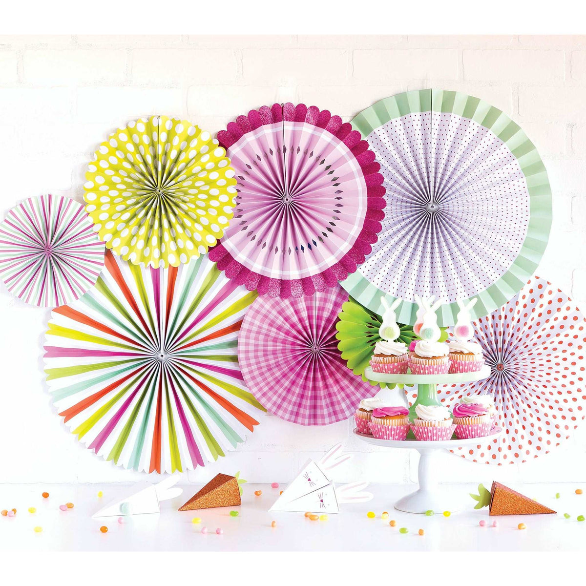 A collection of colorful paper fans in varying pastel and neon patterns in different sizes, displayed against a white wall behid a table with cupcakes with bunny toppers on a white cake stand. carrot and bunny style favor boxes and jelly beans
