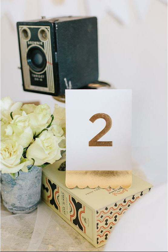 cream tent card with gold foil printed edge and the number 2 on top of a gift box next to a blue vase with white roses and a vintage camera.