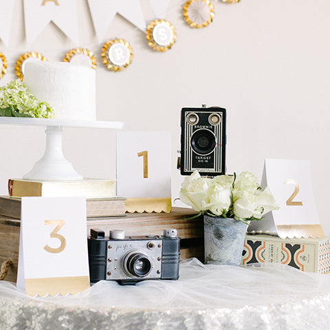 cream tent card with gold foil printed edge and the numbers 2, 1, and 3 surrounded by vintage cameras, a metal vase filled with white roses. white frosted cake on a white cake stand