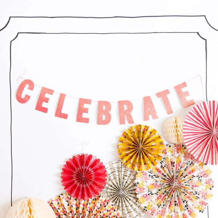Glittery pink letter banner that spells the word celebrate and multi-color printed party fans on a white background 