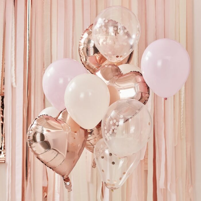 A collection of balloons in rose gold, blush, and peach colors, including round and heart-shaped foil and latex balloons in front of a blush colored tassel style curtain