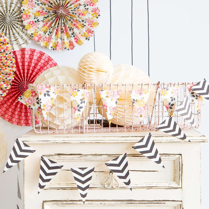 A mini double-sided printed pennant-style banner with a floral print on one side and a classic black and white chevron pattern on the other, with multicolored printed party fans displayed over a rustic wooden cabinet.
