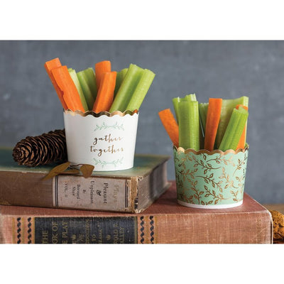 2 stacked vintage books with 1 sage green baking cup with printed gold foil allover floral pattern and rim filled with carrot and celery sticks on the first book, on the second book 1 acorn and 1 white baking cup with printed sage green florals and gold foil words that read gather together