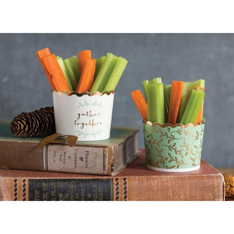 2 stacked vintage books with 1 sage green baking cup with printed gold foil allover floral pattern and rim filled with carrot and celery sticks on the first book, on the second book 1 acorn and 1 white baking cup with printed sage green florals and gold foil words that read gather together