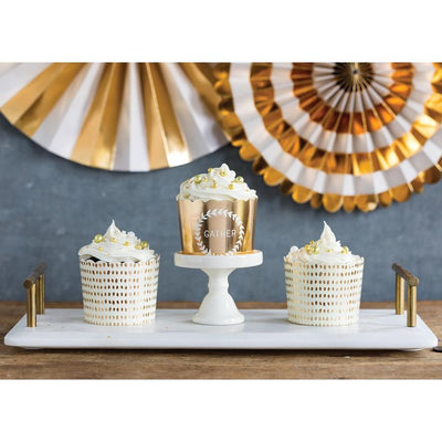 One gold foil and white treat cups with a wreath design and the word 'Gather' in white, and two white treat cups with allover gold foil dot print pattern on a white baking tray, displayed on a table with a festive background.