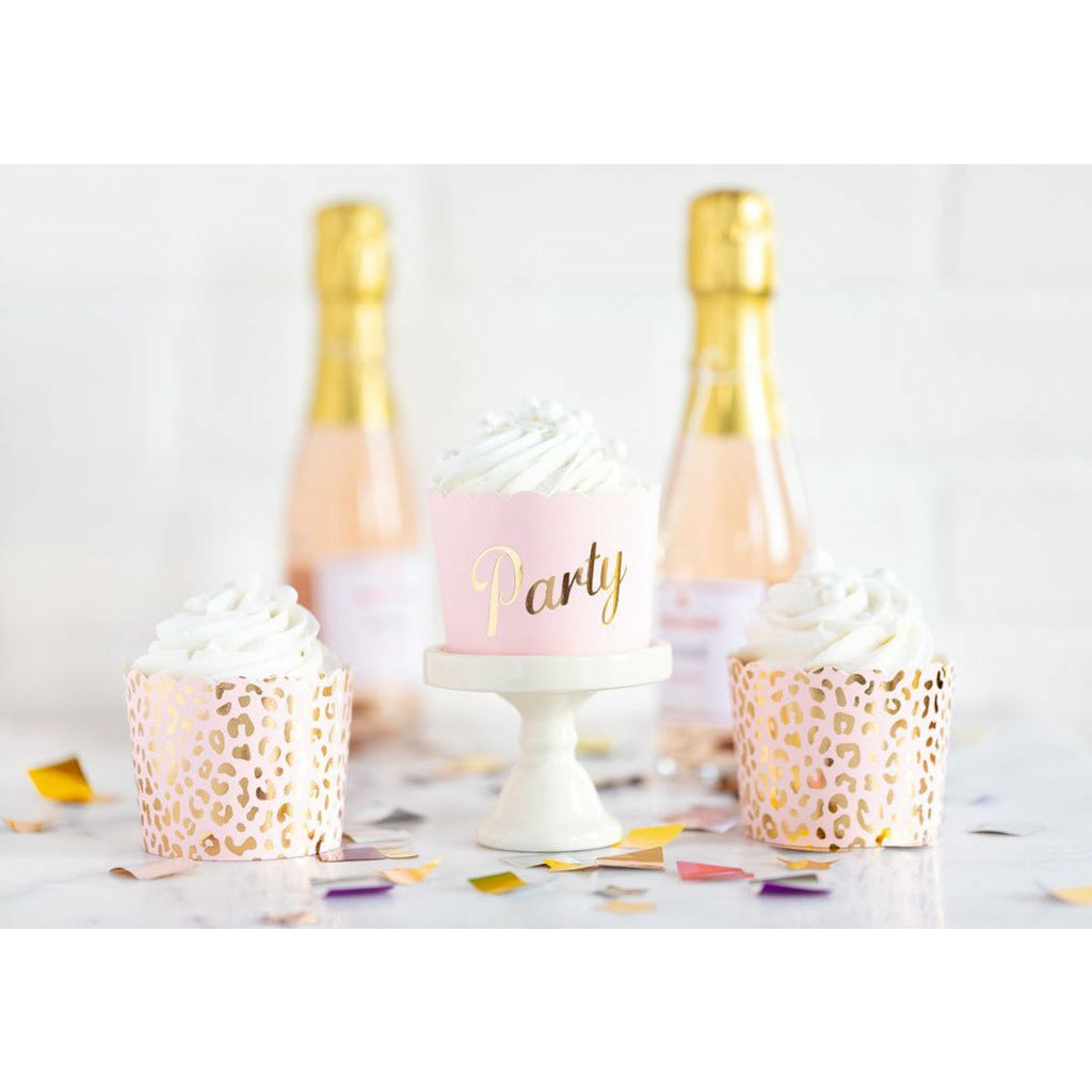 2 champagne bottles, confetti spread on a white table, 2 blush pink food cups with all over gold cheetah print and 1 blush pink food cups with the word party printed in gold on a white cupcake stand