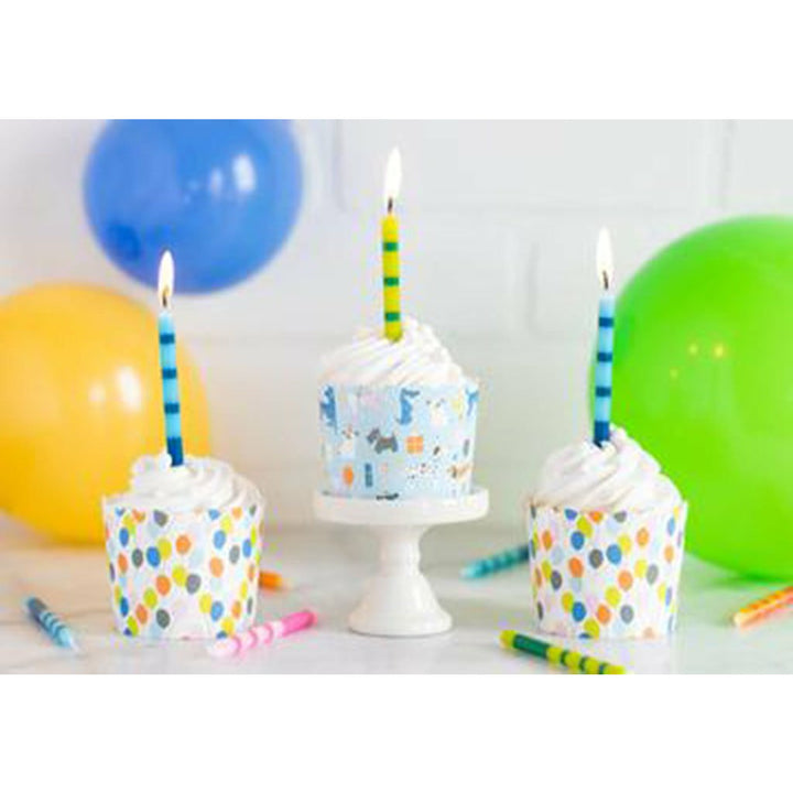 White brick background, a yellow, blue, and green balloon, crayons on a white table. 2 white treat cups with multicolor balloons printed all over and a blue treat cups with various puppies printed all over on a white cupcake stand.