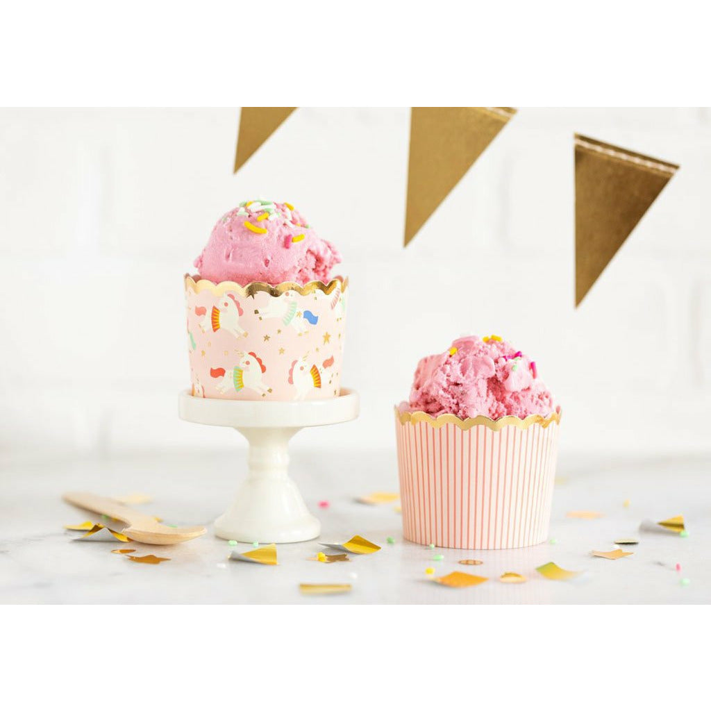 Gold confetti spread across a white table. A 2-tone pink vertical all-over stripe baking cup with strawberry ice cream in it next to a white cupcake stand with a pink gold trimmed baking cup with a unicorn all-over print and strawberry ice cream with sprinkles in it.