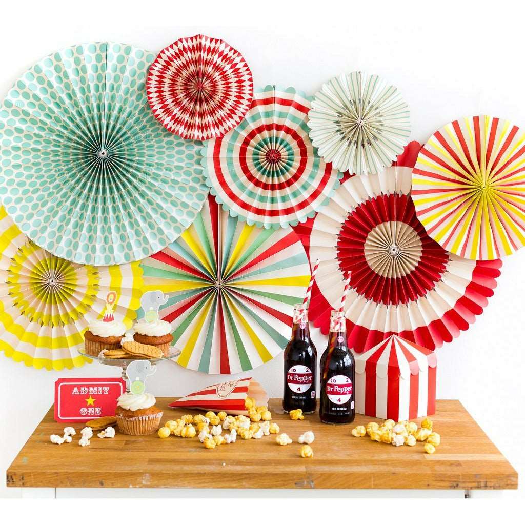 Carnival themed setting with bright colored paper fans on the background wall, 2 vintage Dr Pepper soda bottles, a red and white striped popcorn bag with popcorn scattered on a table. and white frosted cupcakes
