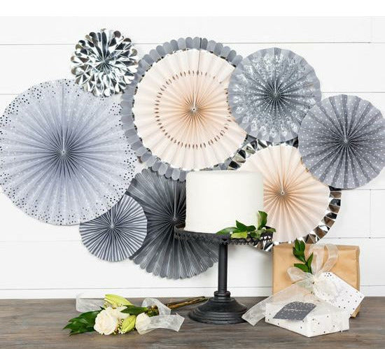 A set of paper fans in grey, silver, and blush pink colors, displayed in various sizes and print patterns on a white wall, a cake on a black cake stand surrounded by gift boxes, and a white rose on a wooden table.