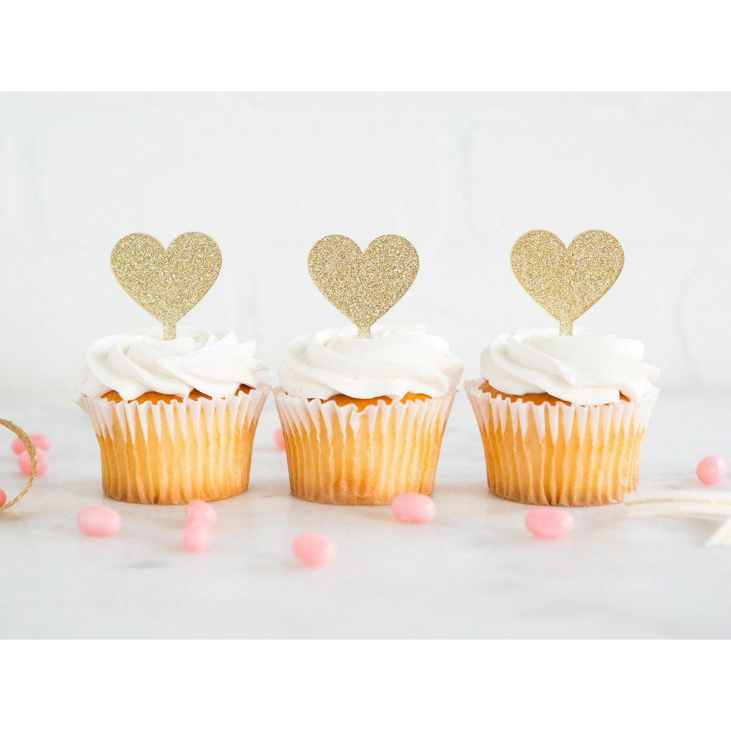 Gold glitter heart-shaped acrylic toppers on cupcakes with pink sprinkles on a white surface.