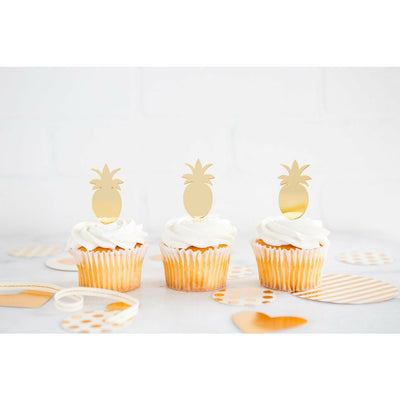 3 gold mirror finish pineapple-shaped cupcake toppers on top of vanilla frosted cupcakes on a white surface with gold and white striped decorations around.