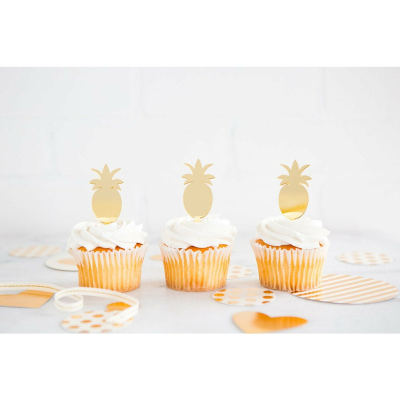 3 gold mirror finish pineapple-shaped cupcake toppers on top of vanilla frosted cupcakes on a white surface with gold and white striped decorations around.