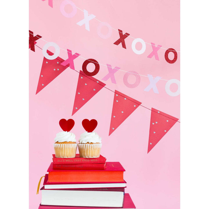 Alternating Red Glitter, Pink, and White Valentine XOXO Letter Banner &amp; Red Pennant with Silver Printed Heart Banner Set hanging above a stack of books with 2 cupcakes on top, each with a red heart-shaped cupcake