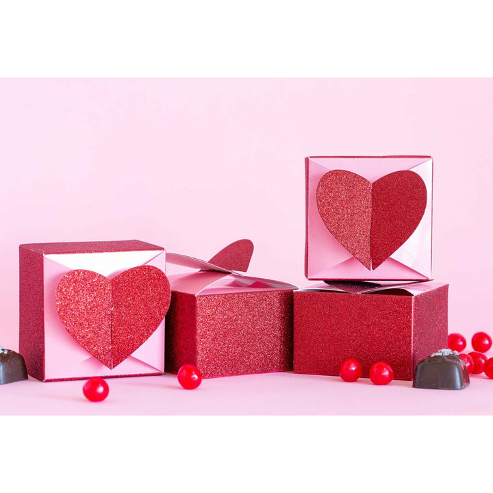 4 Pink and glittery red square favor boxes topped with a 3D glitter red heart-shaped closure. Red jellybeans and chocolate candies are scattered on the table around the boxes.