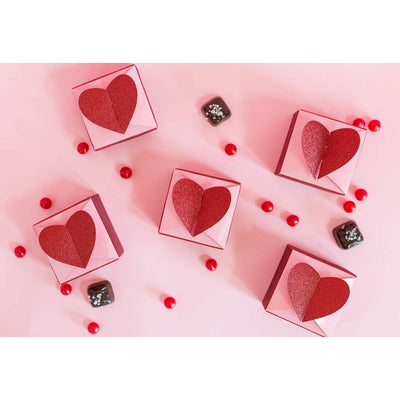 Valentine's Day inspired favor boxes. A top view of five pink and glittery red square favor boxes, topped with a 3D glitter red heart-shaped closure. Red jellybeans and chocolate candies are scattered on the pink colored table around the boxes.