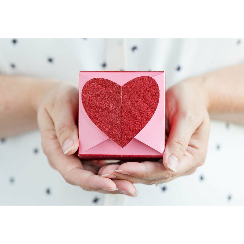 A woman holding a Pink and glittery red square favor box topped with a 3D glitter red heart-shaped closure with both hands. 