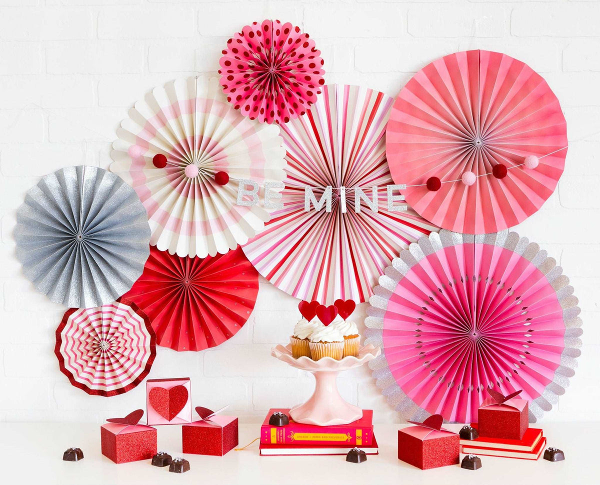A collection of paper fans in various sizes with a Valentine's Day color scheme, including pink, red, cream, and silver on a white brick wall with a red, pink, and silver be mine letter banner. cupcakes topped with red heart toppers, red and pink heart shaped favor boxes, and chocolates on a white table.