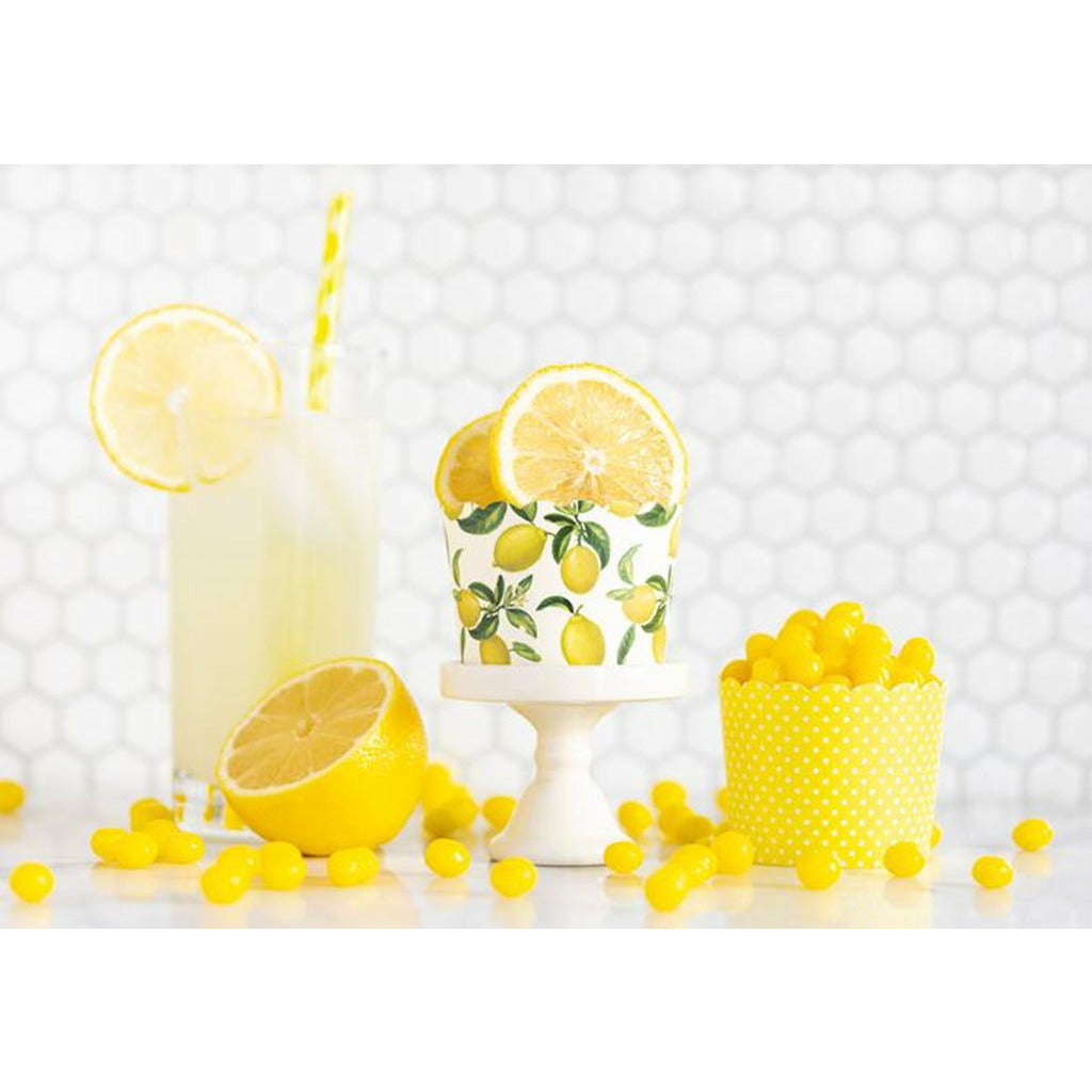 White hexagon tiled background, glass of lemonade with a lemon slice on the rim and a yellow striped straw, a white cupcake stand with a white cupcake cup with all-over printed lemons filled with lemon slices. A yellow cup white polka dot print filled with lemonhead candy.