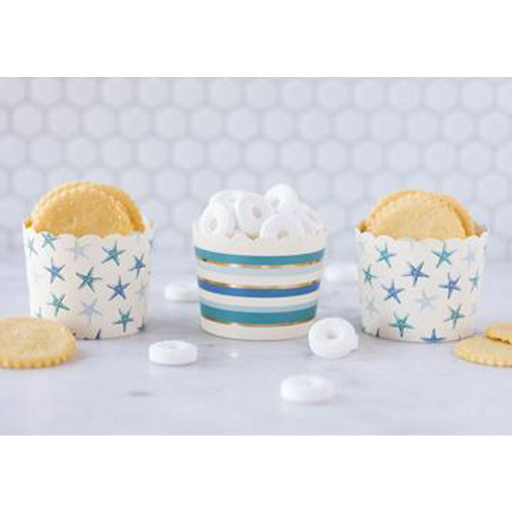 Hexagon tile background with white lifesaver candies and crackers on a white table and 2 cream food cups with all over starfish print in shades of blue and 1 cream food cup with allover stripes in shade of blue and gold foil print.