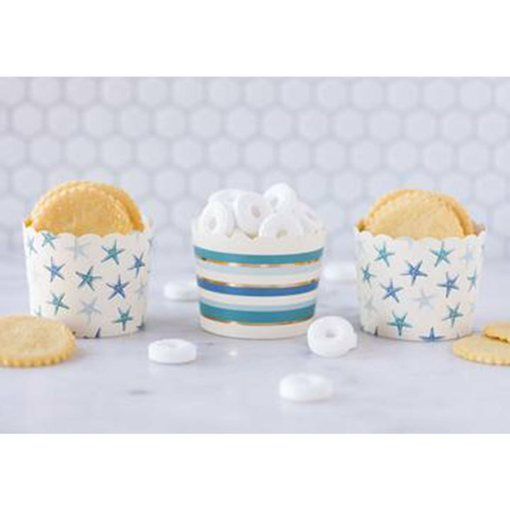 Hexagon tile background with white lifesaver candies and crackers on a white table and 2 cream food cups with all over starfish print in shades of blue and 1 cream food cup with allover stripes in shade of blue and gold foil print.