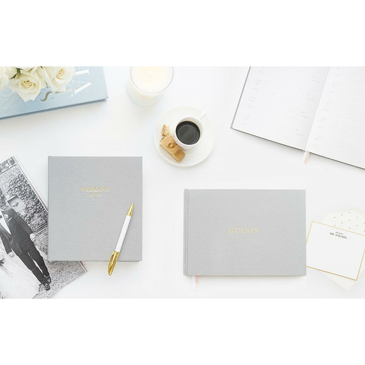 Grey color fabric bookcloth covered guest book with the word guest in gold foil print, next to a grey color fabric bookcloth covered planner book with the words wedding agenda in gold foil print